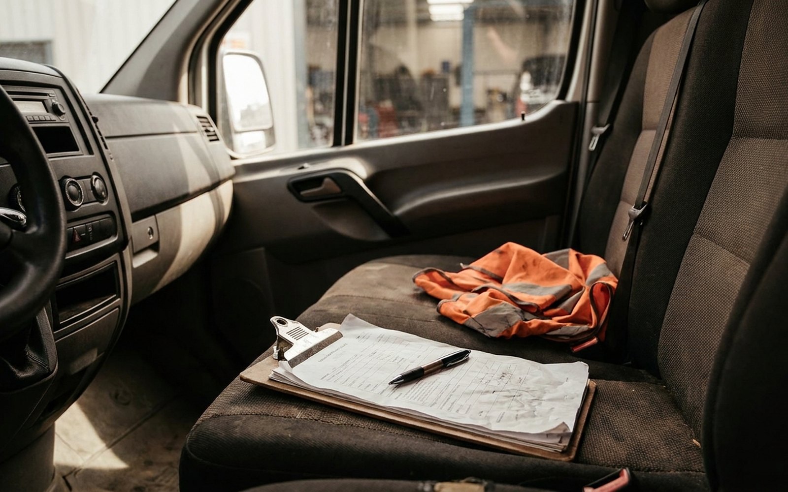 Clipboard with a form and pen on the passenger seat of a work van, an orange safety vest beside it