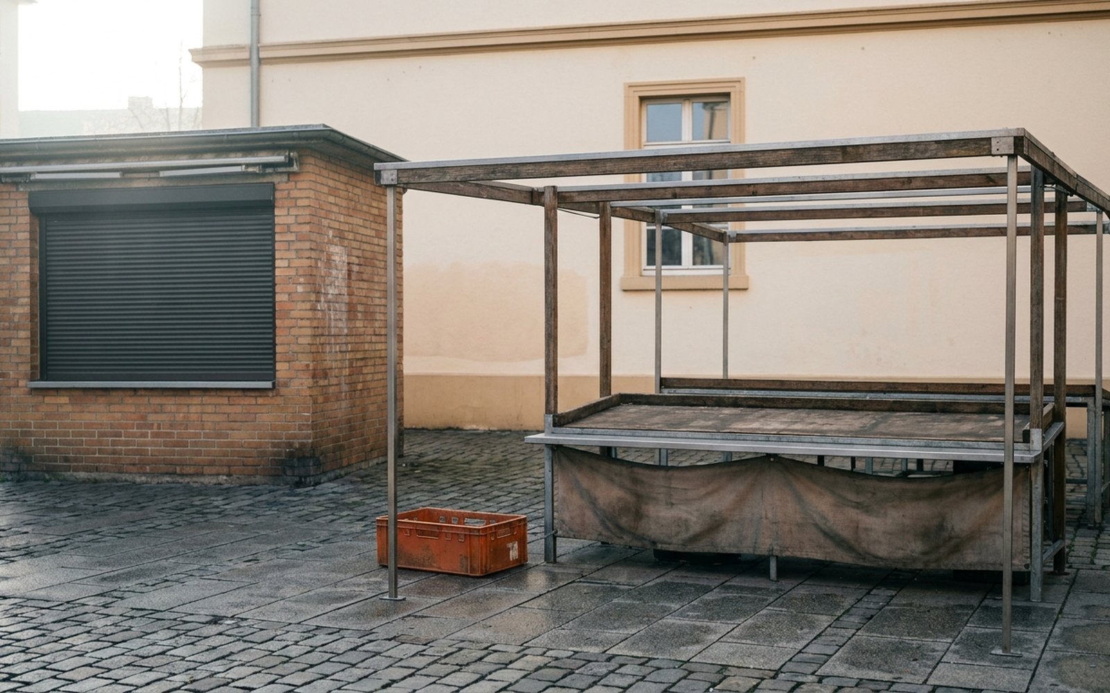 Empty market stall frame next to a permanent kiosk with its shutter down, an orange crate between them